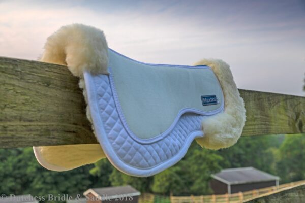 A saddle pad with sheepskin on it hanging from the side of a fence.